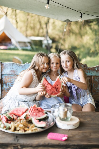 Three cute happy smiling girls, sisters, fiends, sitting at the table on vintage wooden bench and eating watermelon outdoors,