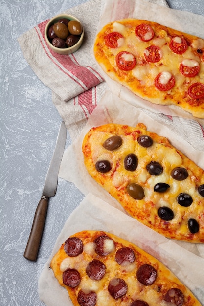 Three different oval pizza with olives, tomato and salami on a gray concrete.   