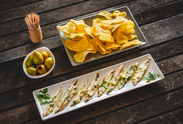 Three dishes with potato chips, picked anchovies and green olives on a dark wooden table with toothpicks. Typical Spanish snacks.