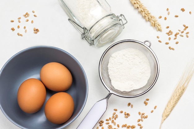 Three eggs in gray bowl Flour in sieve and glass jar Milk and spikelets of wheat