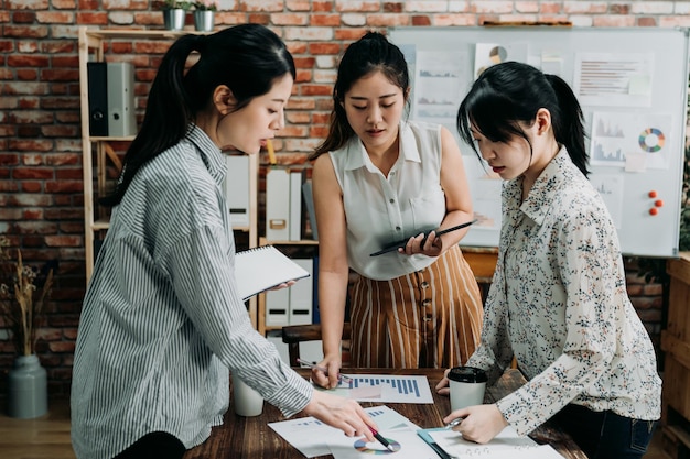 会話をし、テクノロジーを使用しているオフィスの3人の女性ビジネスマン。忙しい職場の机に立って、新しいプロジェクトについて話し合っている若い創造的なチームワークパートナーの女性のグループ。