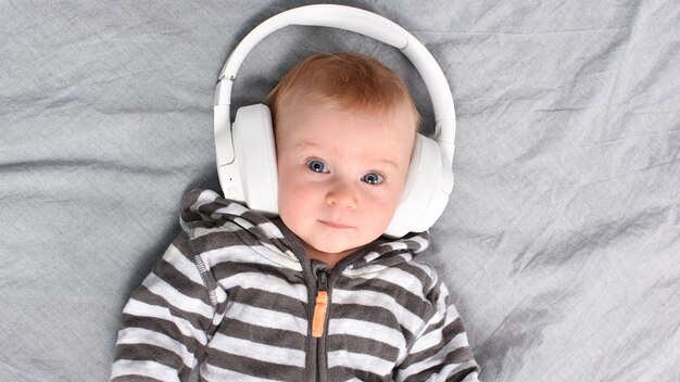 Photo three-four months old cute caucasian baby lying on bed listening to music in big headphones.