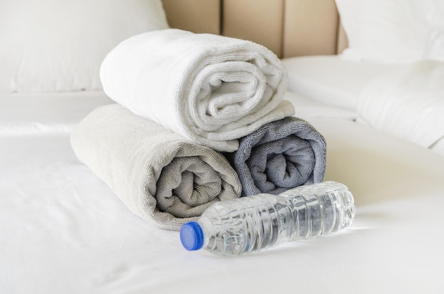 Three fresh clean bath towels rolled up in rolls and bottle of water on a white bed in hotel room after cleaning