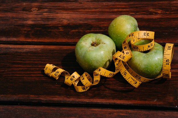Три свежих зеленых яблока лежат в ряд на деревянном столе и обмотаны желтой измерительной лентой