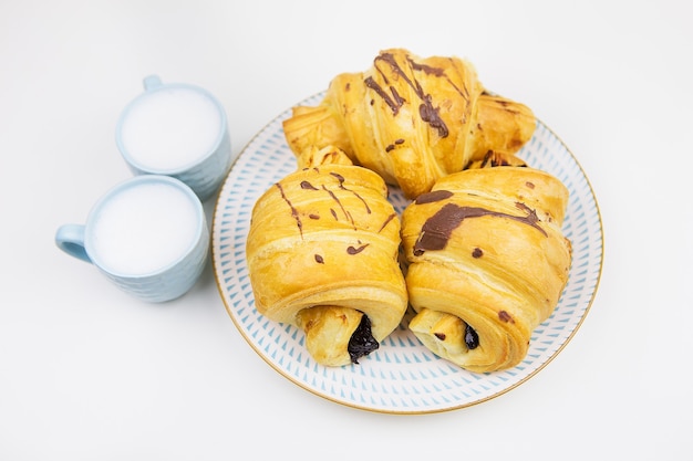 Three freshly baked croissants on a white ceramic plate, two cups of coffee