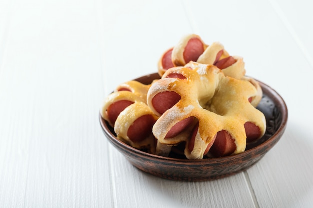 Three freshly cooked homemade sausage rolls in the shape of an asterisk in a clay bowl on a white rustic table. Tasty meat snack made of dough and sausages.