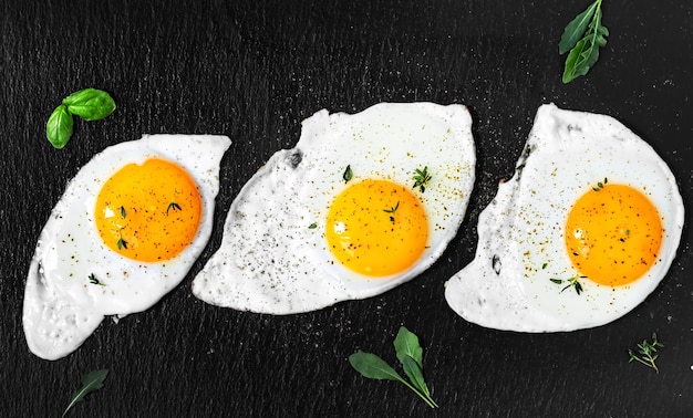 Three fried eggs on a black stone pan with pepper, arugula and basil leaves