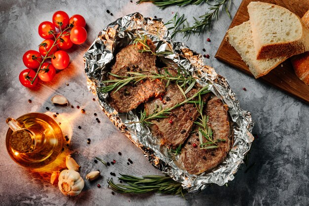 Three fried slices of beef steak in foil with rosemary, cherry tomatoes, slices of bread and olive oil.