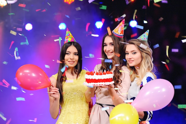 Three girls in festive hats and balloons and cake in hand. 