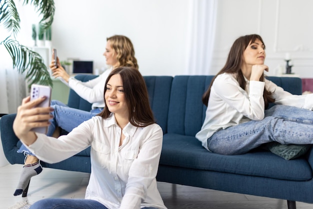 Three girls friends in shirts and jeans sit on and near the sofa in the room and climb social networks