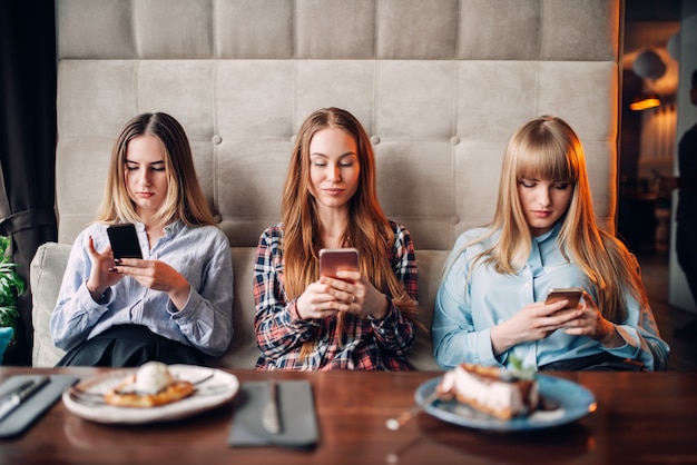 Tre ragazze sedute sul divano e utilizzando i telefoni cellulari nella caffetteria. dessert al cioccolato e alcol sul tavolo