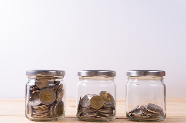 Photo three glass jars with coins inside on wooden table and white wall