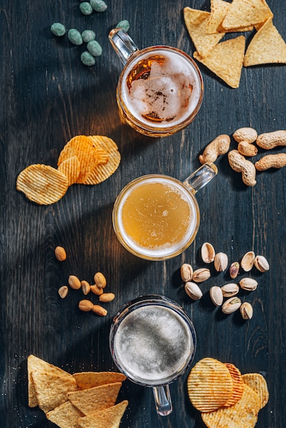 Three glasses of expensive craft beer, classic and unfiltered and dark in a glass on the table with a snack of peanut and pistachio chips and nachos