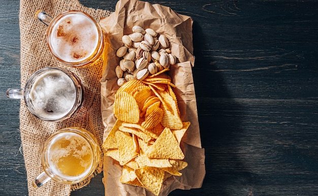 Tre bicchieri di costosa birra artigianale, classica e non filtrata e scura in un bicchiere sul tavolo con uno spuntino di patatine e nachos di arachidi e pistacchi