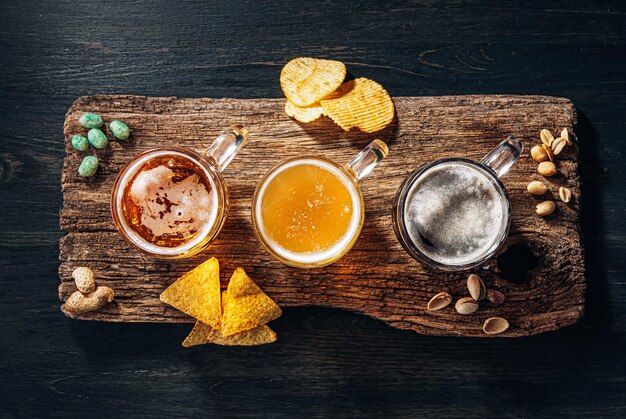 Tre bicchieri di costosa birra artigianale, classica e non filtrata e scura in un bicchiere sul tavolo con uno spuntino di patatine e nachos di arachidi e pistacchi