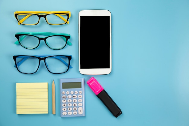 Three glasses yellow notebook mobile phone calculator and hilight marker on blue background pastel style with copyspace flatlay clipping path on screen moblie