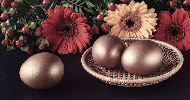 Three golden Easter eggs in rattan plate on dark background with Springtime flowers, herbera daisy. Reflections in metal tray.