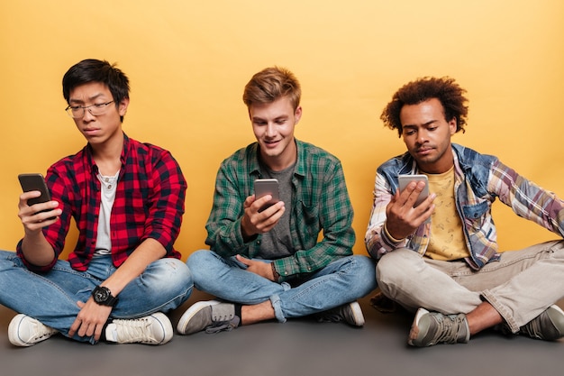 Photo three handsome young men friends using cell phone over yellow background