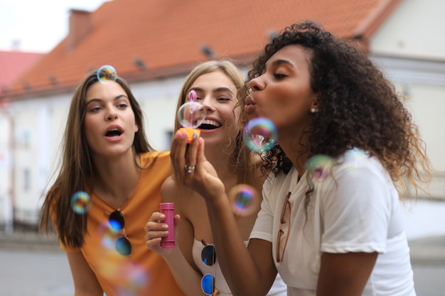 Three happy beautiful women blowing soap bubbles in city street.