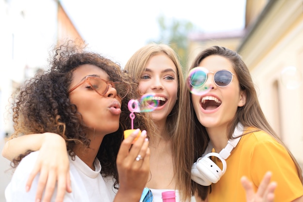 Three happy beautiful women blowing soap bubbles in city street.