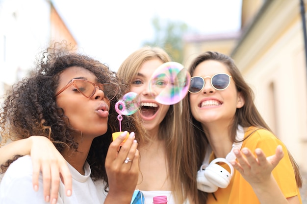 Three happy beautiful women blowing soap bubbles in city street.