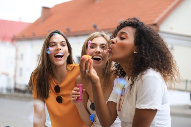 Photo three happy beautiful women blowing soap bubbles in city street.