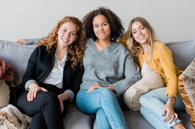 Three happy casual girls sitting on couch at home while enjoying party or leisure in cozy environment