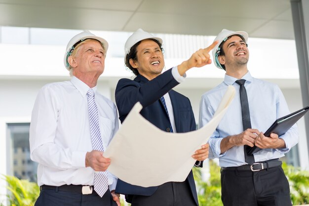 three happy diverse business people wearing helmets, holding blueprint