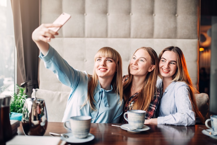 Фотография три счастливые подруги делают селфи на камеру телефона в кафе. подруги сидят в ресторане и веселятся, встречаются сплетники