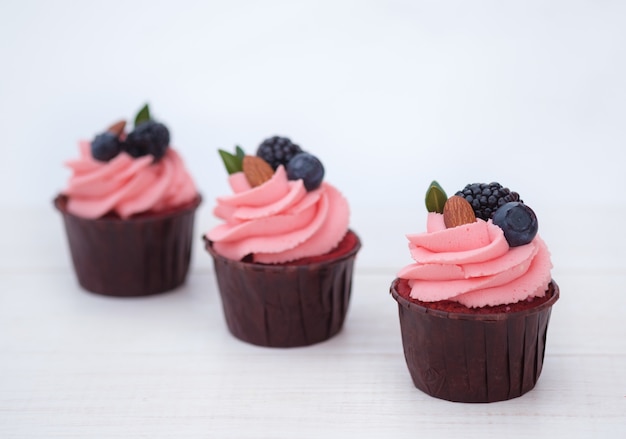 Three homemade cupcakes with pink cream, berries and almonds, on white background