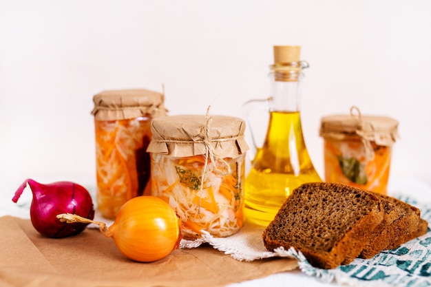 Three jars of sauerkraut and carrots in its own juice with spices and a bottle of oil, bread and onion, white wooden table. traditional home-made