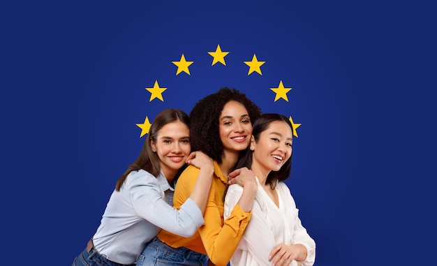 Tre donne gioiose di diverse etnie che si abbracciano e sorridono davanti a una bandiera dell'unione europea