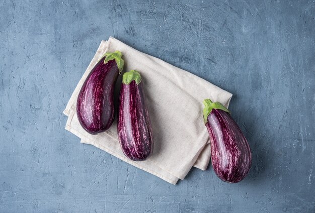 Three  juicy eggplants graffiti  lie on a gray napkin on  blue concrete table top view copy space village