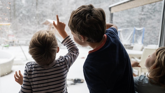 Three kids brothers and sister looking out the window pointing to beautiful winter nature