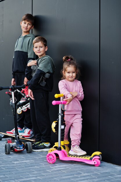 Three kids outdoor against black modern wall. Sports family spend free time outdoors with scooters and skates.