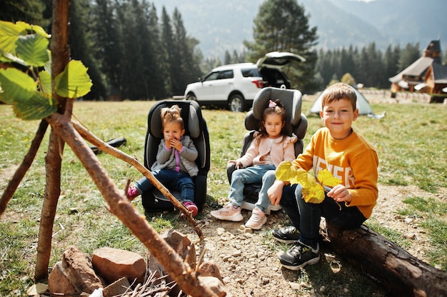 Three kids sitting by the bonfire in mountain. Autumn hike weather. Warming and cooking near flame together.