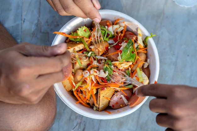Три мужские руки с вилками едят тайский овощной салат вместе с сосисками, вид сверху, крупным планом