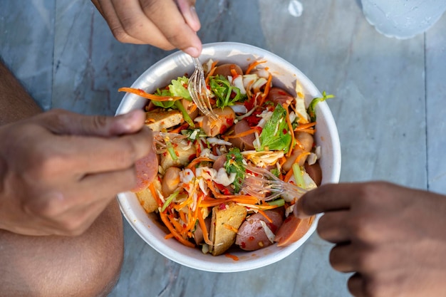 Три мужские руки с вилками едят тайский овощной салат вместе с сосисками крупным планом