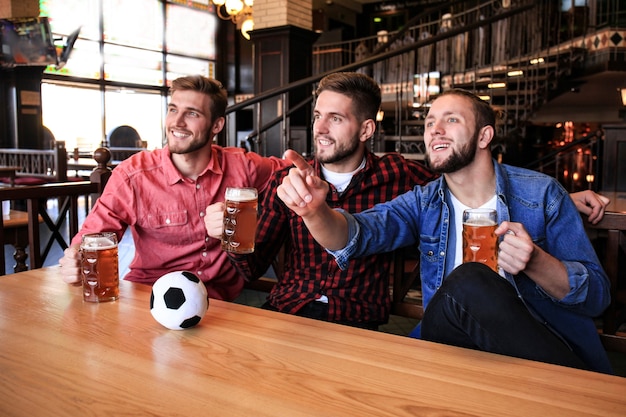 Three men in casual clothes are cheering for football and holding bottles of beer while sitting at bar counter in pub.
