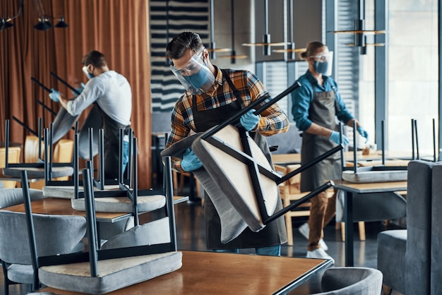 Three men in protective face shields and aprons arranging furniture in restaurant