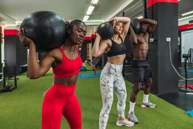 Three multiracial bodybuilder athletes doing toning exercises with weight ball in a gym. Two African bodybuilders and a Caucasian bodybuilder doing crossfit workout routine.