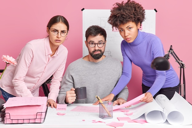 Three multiracial engineers examine draft and talk with each other seriously work on architectural project in contemporary office pose at desktop. Experienced workers check sketch of young employee