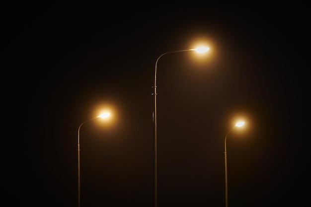 Three night lamppost shines with faint mysterious yellow light\
through evening fog at quiet night