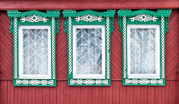 Фото Три старых окна с решетками и резными деревянными зелено-белыми архитетравами на красной стене из доски