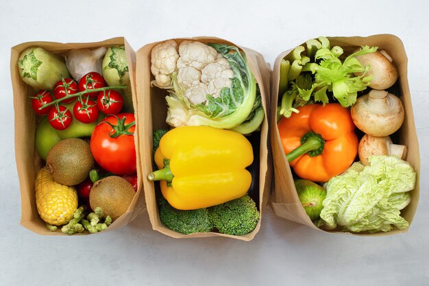 Three paper bags with fresh vegetables and fruits on a light table, top view