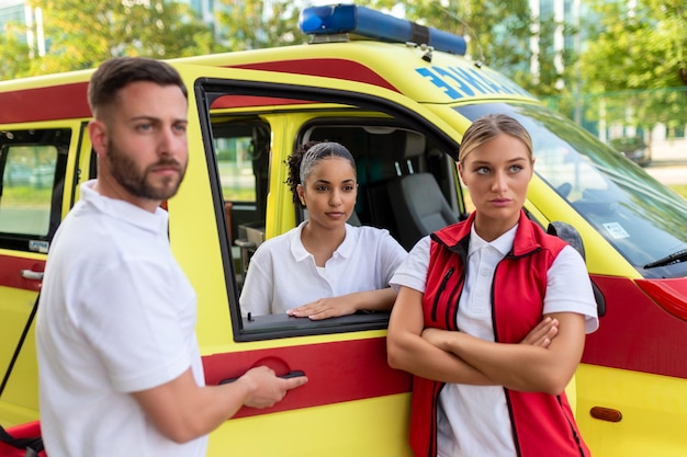 Три фельдшера стоят у машины скорой помощи и разговаривают Парамедик-мужчина с сумкой для медицинских травм Группа из трех фельдшеров, стоящих перед машиной скорой помощи с улыбкой