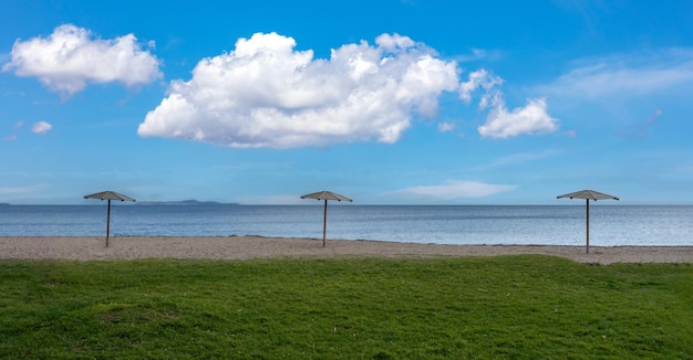 Three parasol on empty sandy and green grass beach Winter sea blue sky background Greek seascape