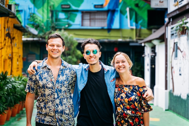 Three people of different ethnicities hugging each other behind the back in the middle of a street with colored facades decorated with street art