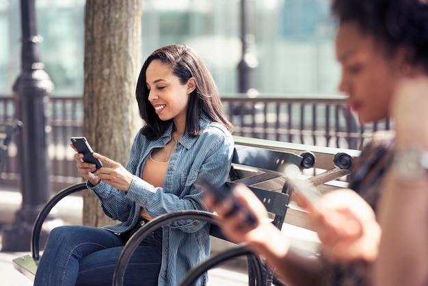 Three people seated side by side on a park bench checking their smart phones