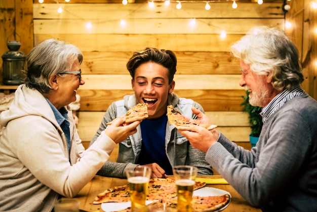 Three people at the table with pizza - two seniors trying to doing eat at the teenager pizza - celebrating new year or some party together